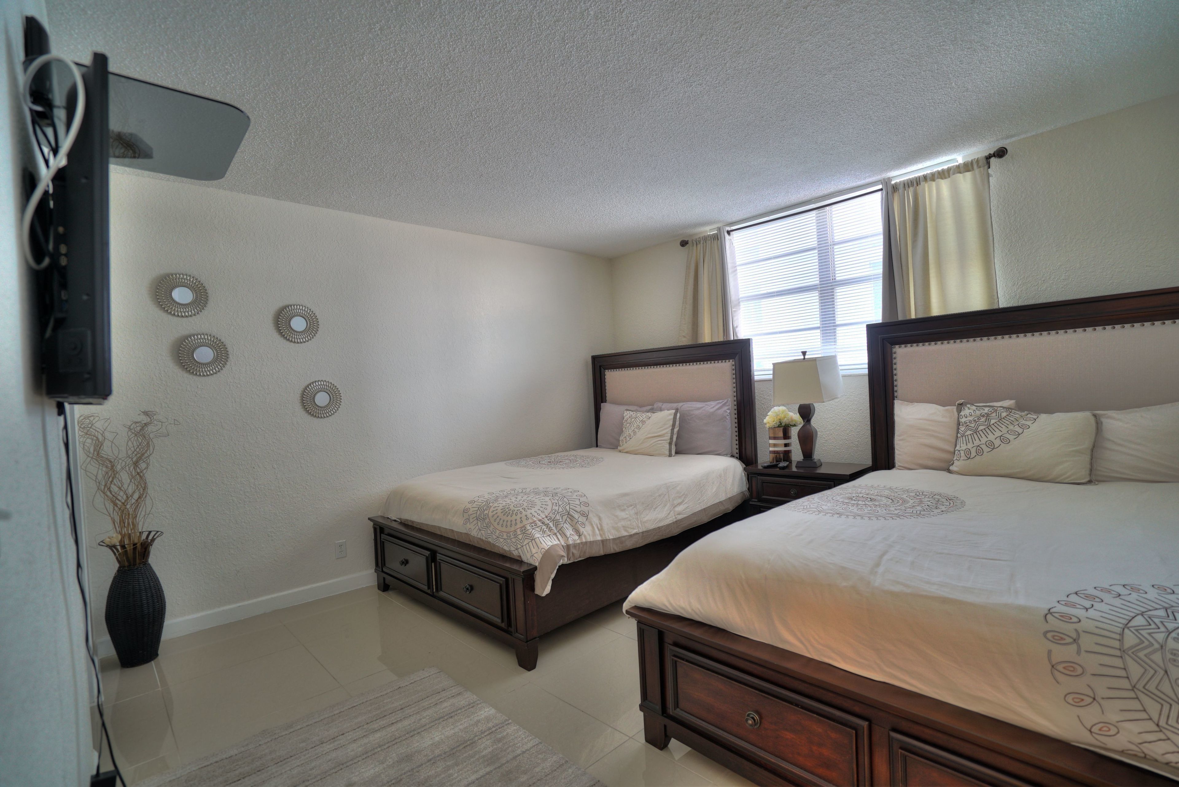 A bedroom with two beds, a bedside table with a lamp, a vase with dried plants, and a wall-mounted TV.