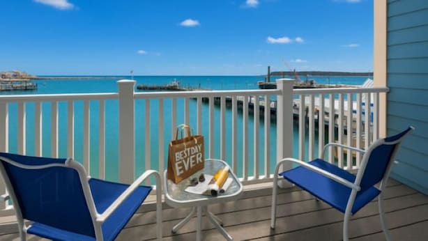 A balcony with two blue chairs, a small table with a 'Best Day Ever' sign, and a view of the ocean and pier. - Bicycle Street Inn (Mackinac Island)