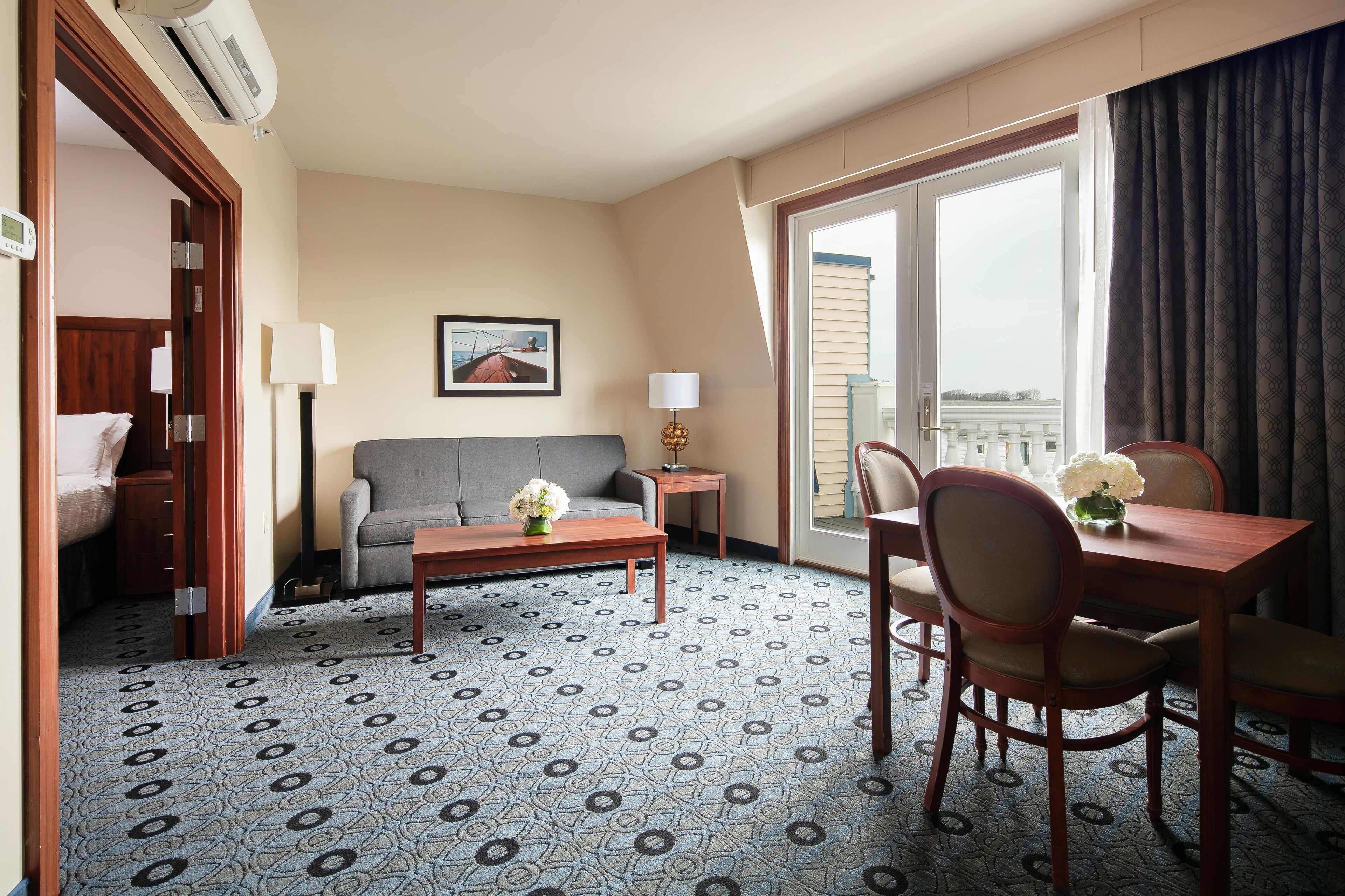 A hotel room with a patterned carpet, a sofa, a dining table with chairs, a small side table, a lamp, and a large window with curtains.