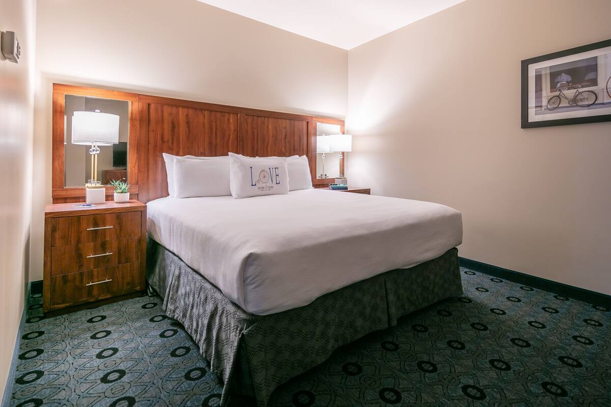 A hotel room with a large bed, wooden headboard, two bedside tables with lamps, a mirror, and a framed picture on the wall.