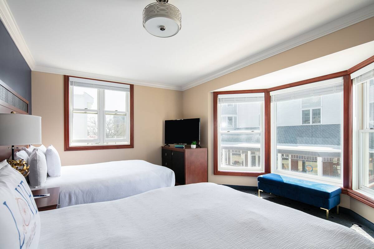 A hotel room with two beds, a TV, a bench, and large windows offering a view of the city.