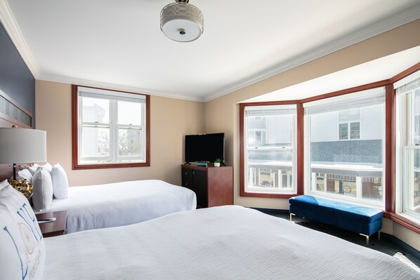 A hotel room with two beds, a TV, a bench, and large windows offering a view of the city.