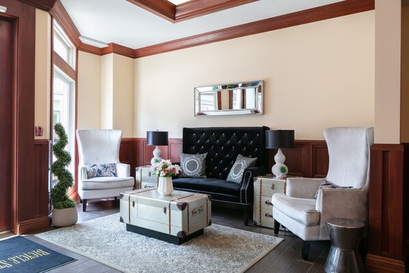 A living room with a black tufted sofa, white armchairs, a white coffee table with a vintage suitcase, and a mirror on the wall. - Bicycle Street Inn (Mackinac Island)