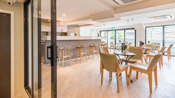 A modern dining area with wooden chairs and tables, a glass partition, and a kitchen in the background.