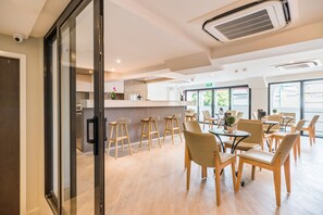 Un comedor moderno con sillas y mesas de madera, un separador de vidrio y una cocina al fondo.