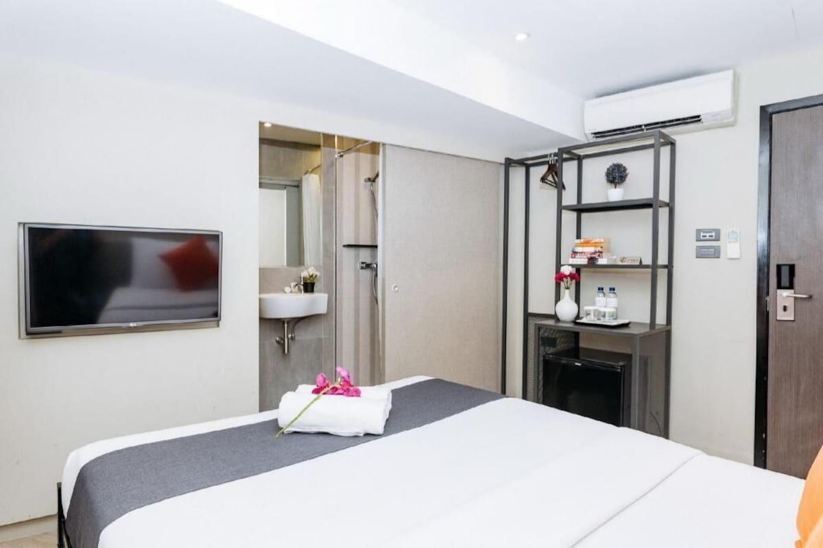 Habitación de hotel con cama, televisión, ducha, lavamanos y estante con artículos de tocador.