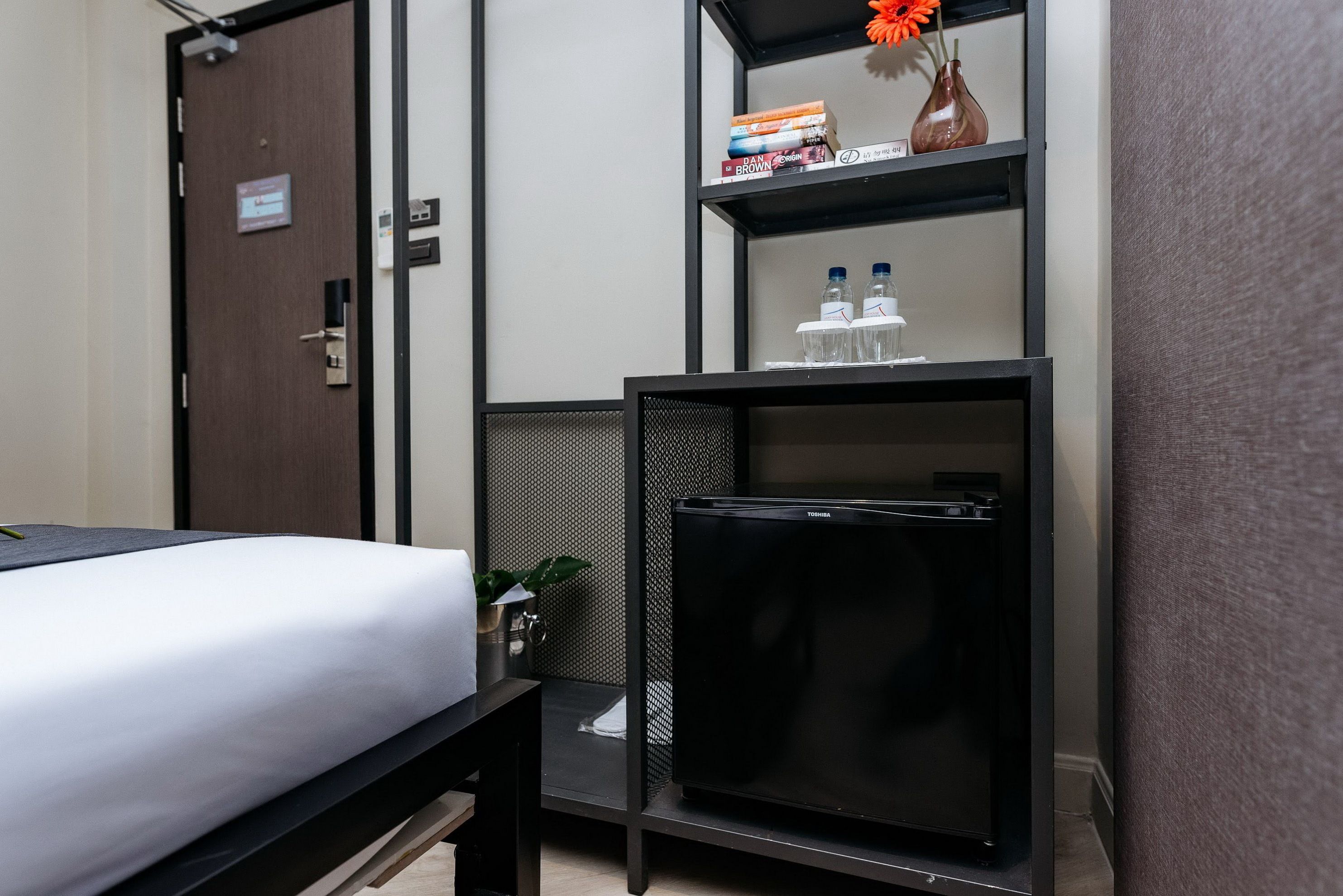 Une chambre d’hôtel avec un lit, un bureau, un meuble noir contenant des livres et un vase, et une porte avec une pancarte.