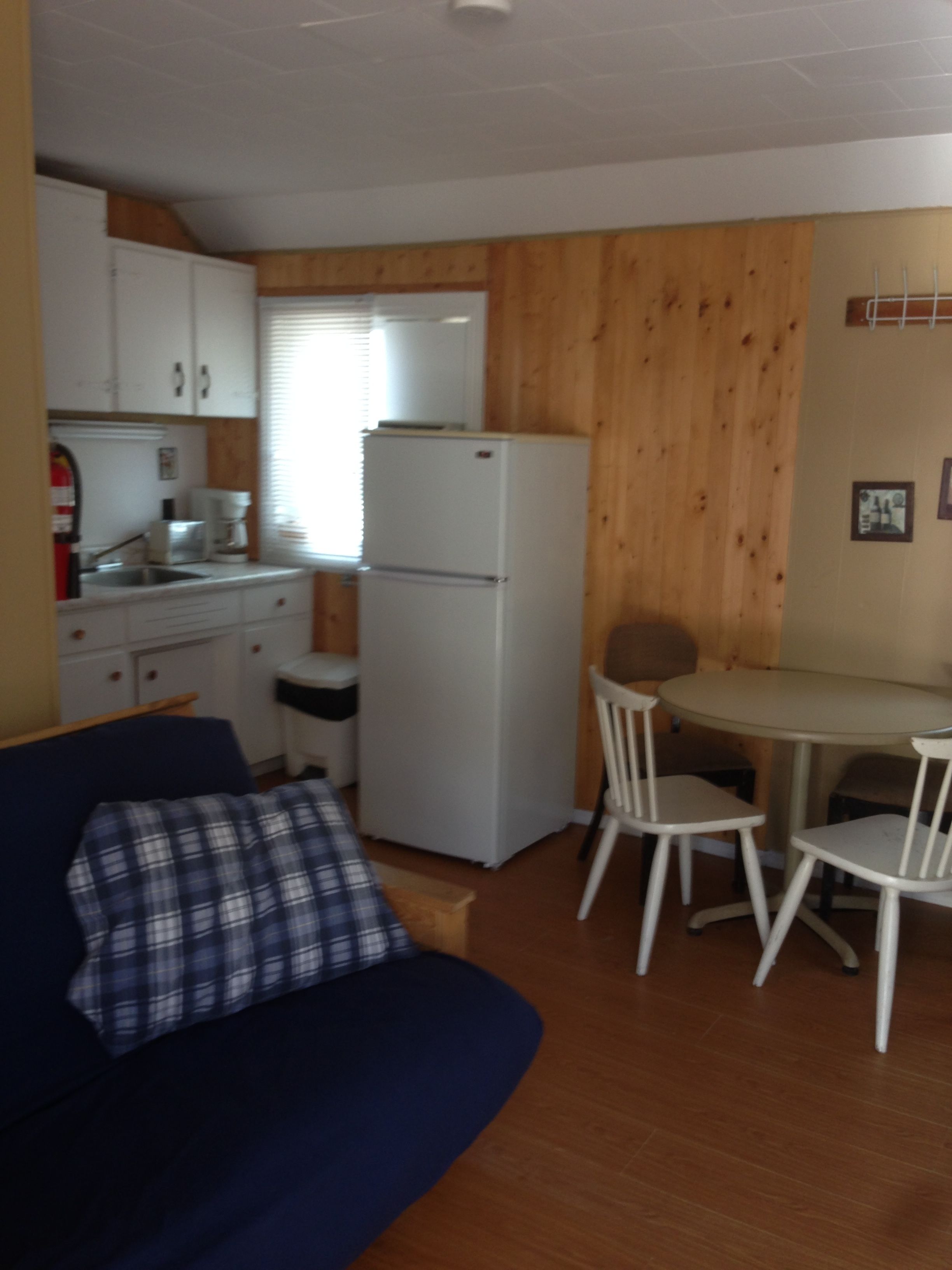 A compact kitchen with white cabinets, a white refrigerator, a coffee maker, and a sink. There is a blue sofa with a plaid pillow in the foreground.
