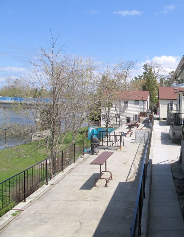A riverside walkway with picnic tables and benches, a small bridge, and houses in the background. - Edgewater Resorts - Edgewater Cottages (Wasaga Beach)