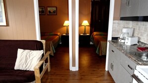 Habitación de hotel con sofá de madera, dos camas individuales, una mesita con lámpara, kitchenette con cafetera y tostadora, y un baño visible a través de una puerta abierta.