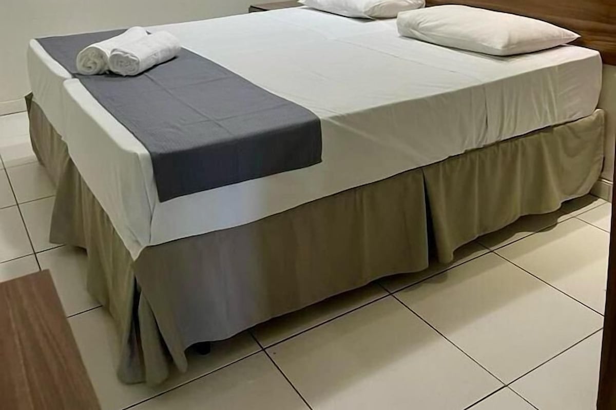 Una cama doble bien hecha, con colcha gris y almohadas blancas, cabecero de madera y una ventana con persianas.