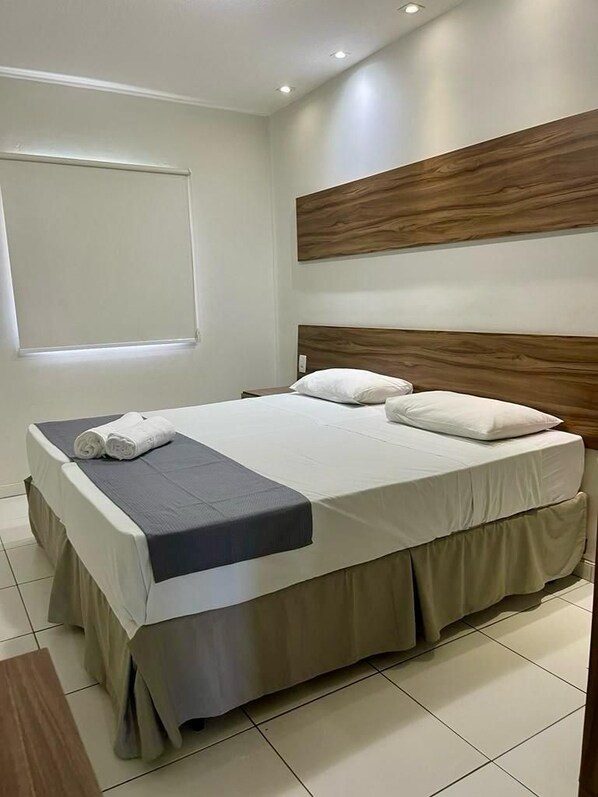 A neatly made bed with white linens and a dark blue bedspread, a wooden headboard, and a window with blinds. - Hotel Pousada Alagoana (Maceió)