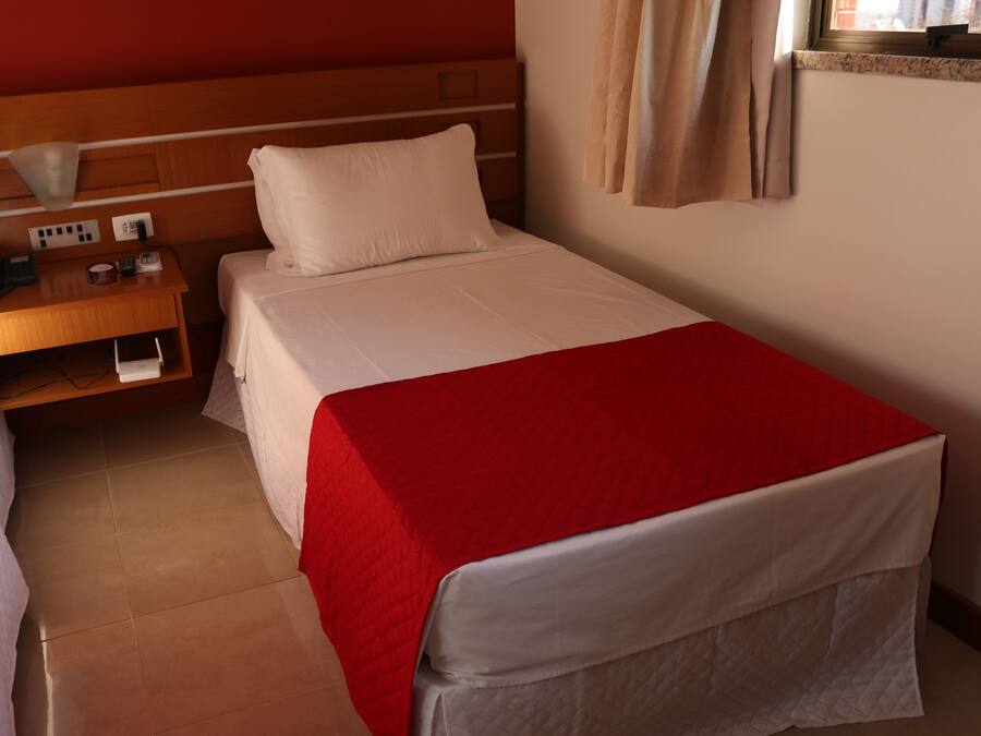 Una habitación de hotel con una cama individual, una mesita de noche de madera con lámpara y una ventana con cortinas.