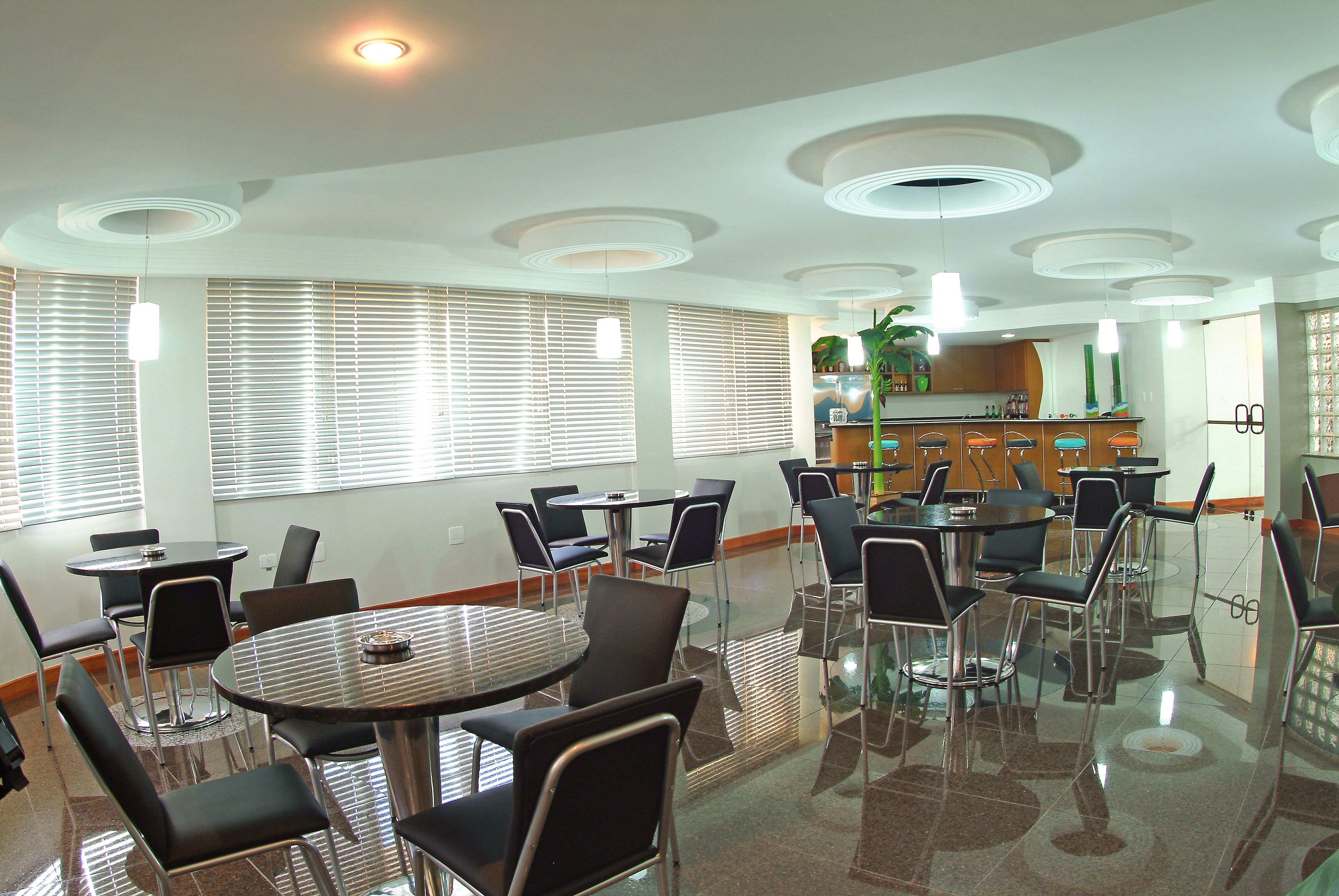 Un comedor moderno con mesas y sillas redondas, ventanas amplias con persianas y una cocina al fondo.