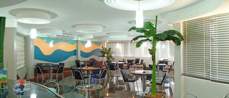 Un espace repas moderne avec des tables et des chaises rondes, une grande plante et une fresque colorée sur le mur.