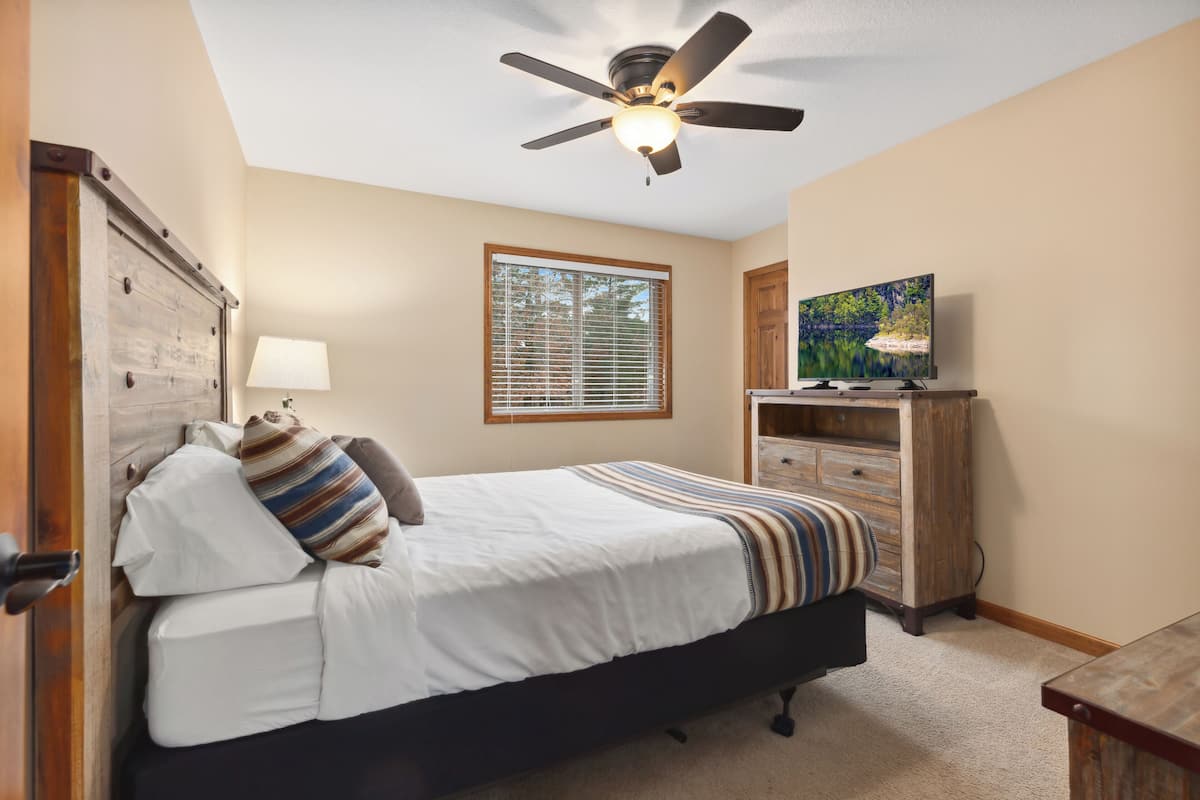 A bedroom with a bed, a ceiling fan, a window with blinds, a TV on a wooden stand, and a dresser.