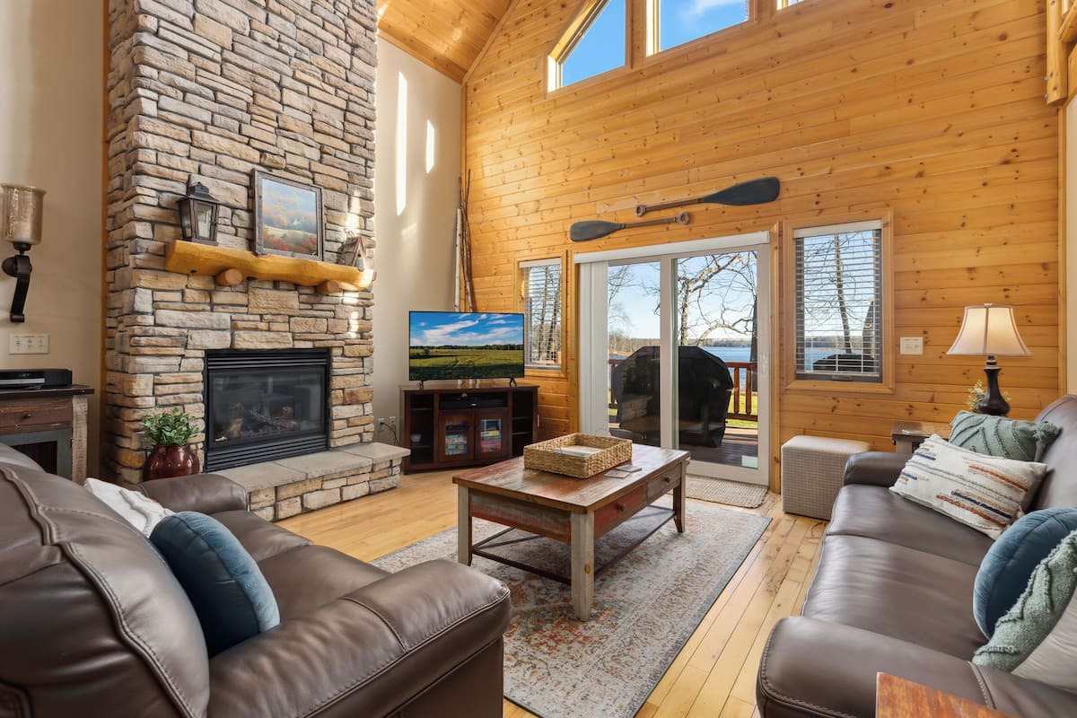 A cozy cabin interior with a stone fireplace, wooden walls, a large window, and a comfortable seating area.