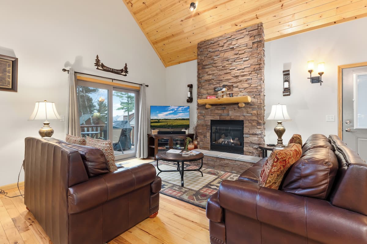 A cozy living room with leather sofas, a stone fireplace, a television, and a large window overlooking a deck.