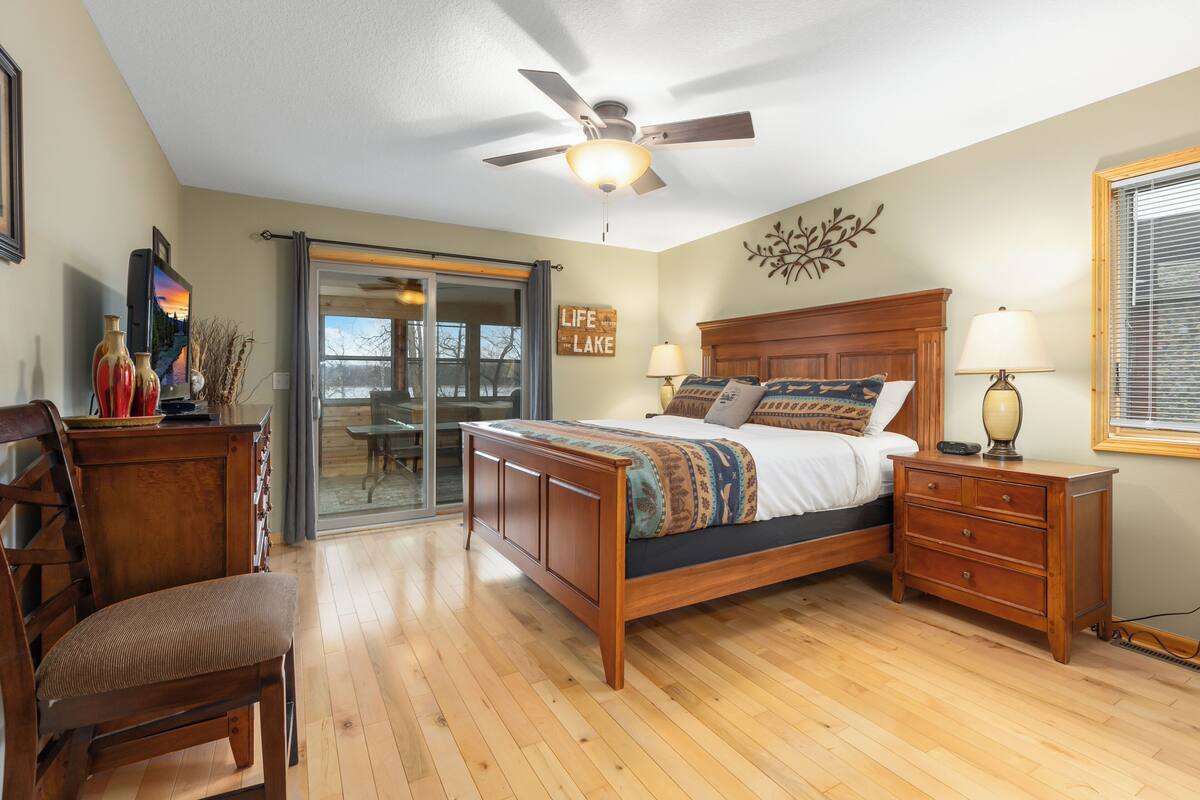 A bedroom with a wooden bed, a nightstand, a chair, a ceiling fan, and a view of a lake through a sliding door.