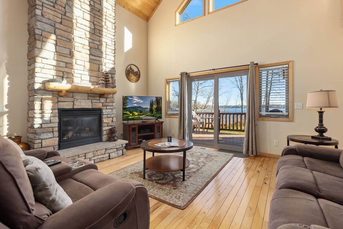 A spacious living room with a stone fireplace, a large TV, and a view of a deck and lake.