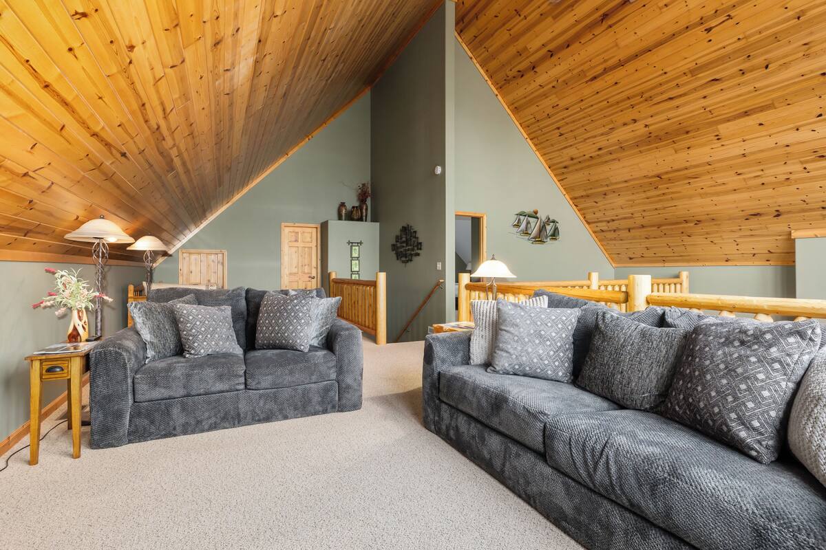 A cozy living room with two grey sofas, wooden ceiling beams, and a staircase leading to an upper level.
