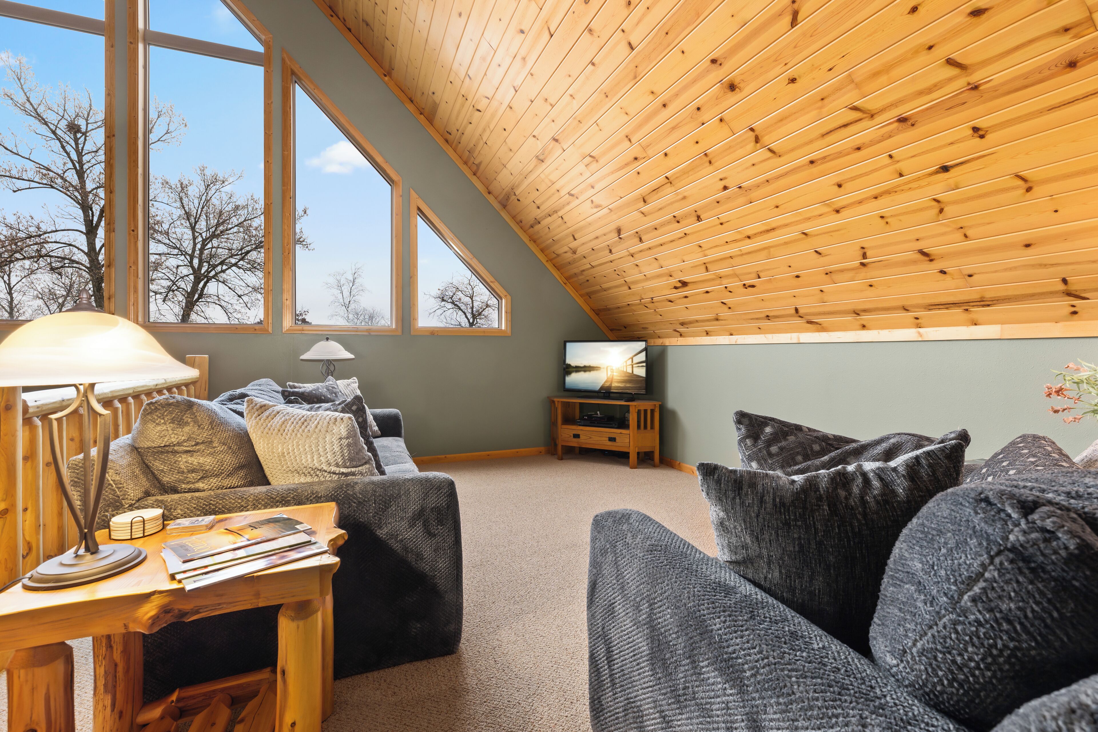 A cozy living room with a sloped ceiling, large windows, a television, and a comfortable seating area.