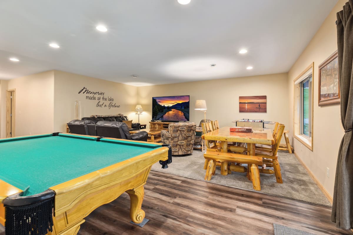 A pool table, a sofa, a wooden dining table, and chairs in a room with wooden flooring and beige walls.