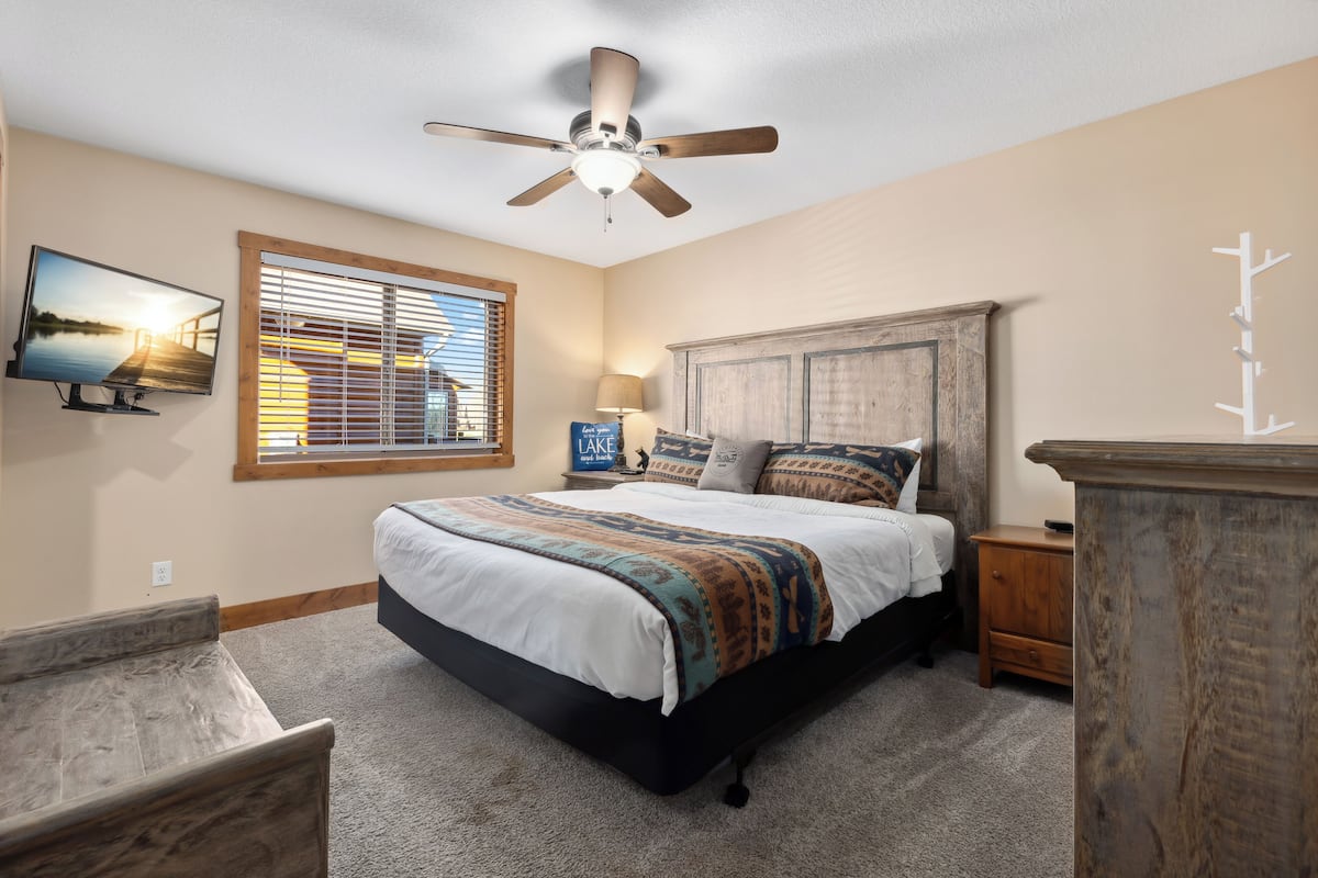 A bedroom with a bed, a ceiling fan, a TV, a window with blinds, a nightstand, and a dresser.
