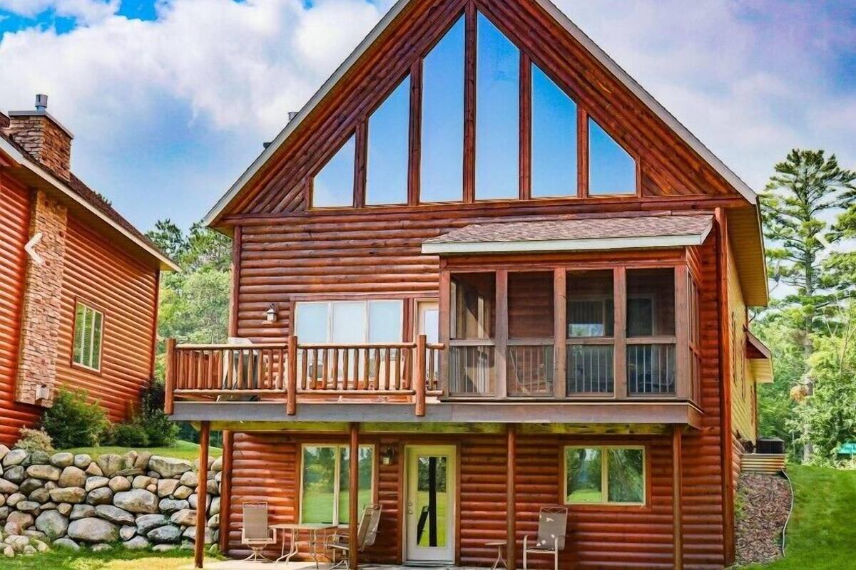 A two-story log cabin with a balcony, stone foundation, and a stone wall.