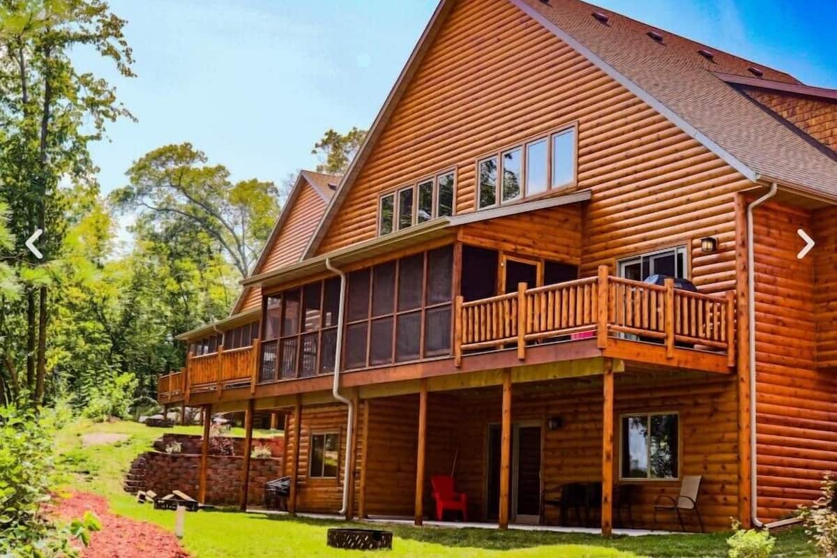 A two-story log cabin with a balcony, large windows, and a covered porch area.
