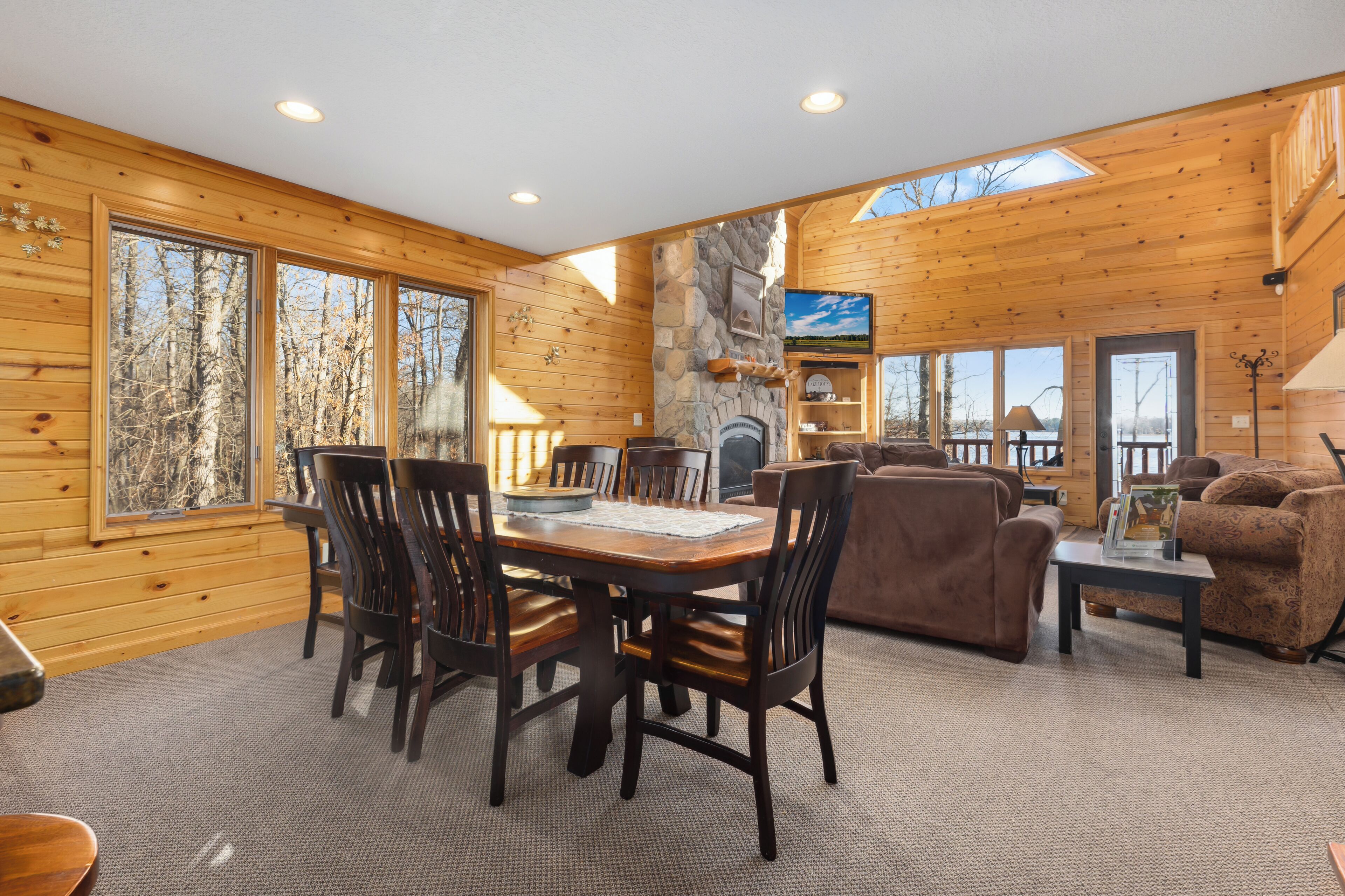 A wooden cabin interior with a dining table, chairs, a fireplace, a sofa, and large windows.