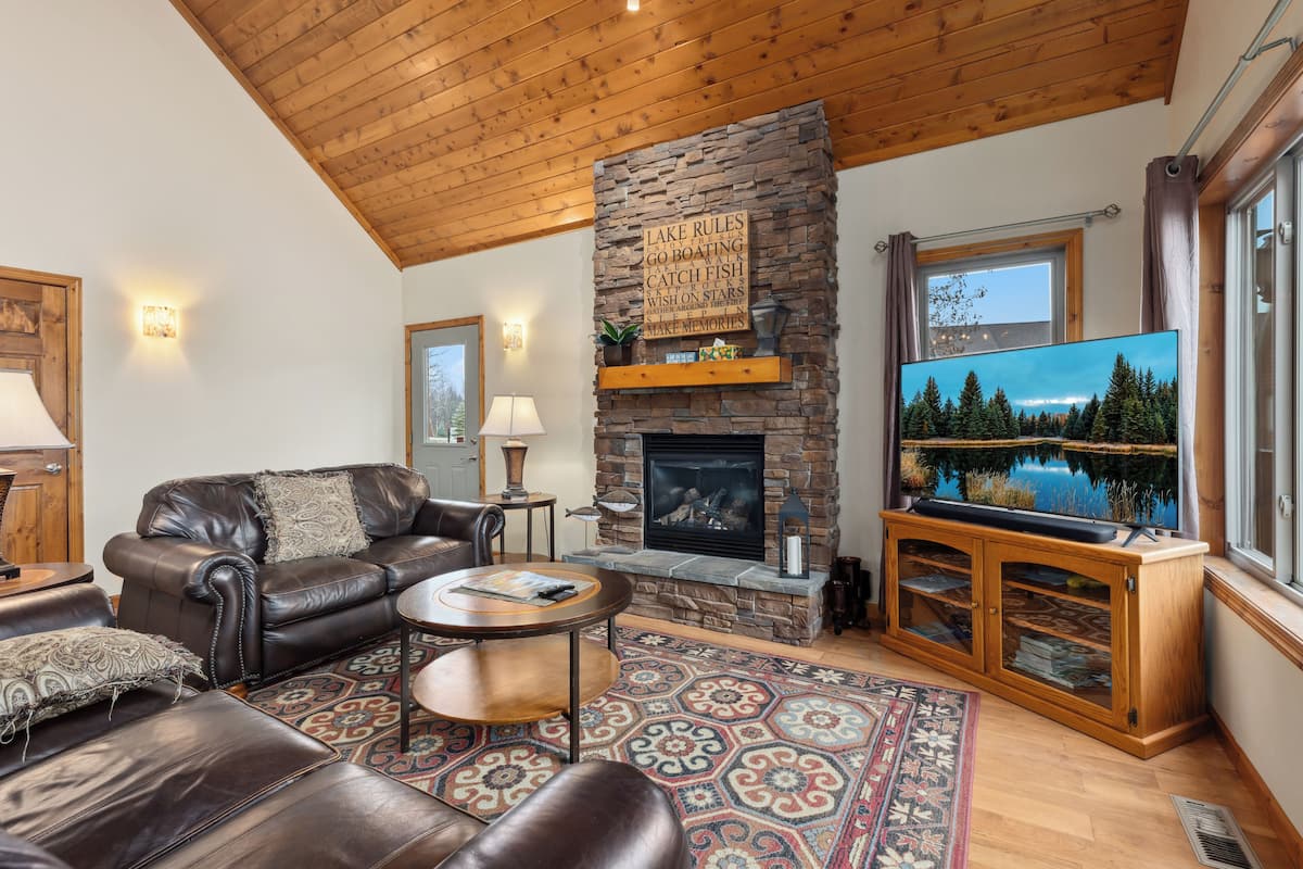 A cozy living room with a stone fireplace, leather furniture, and a large TV screen displaying a scenic view.