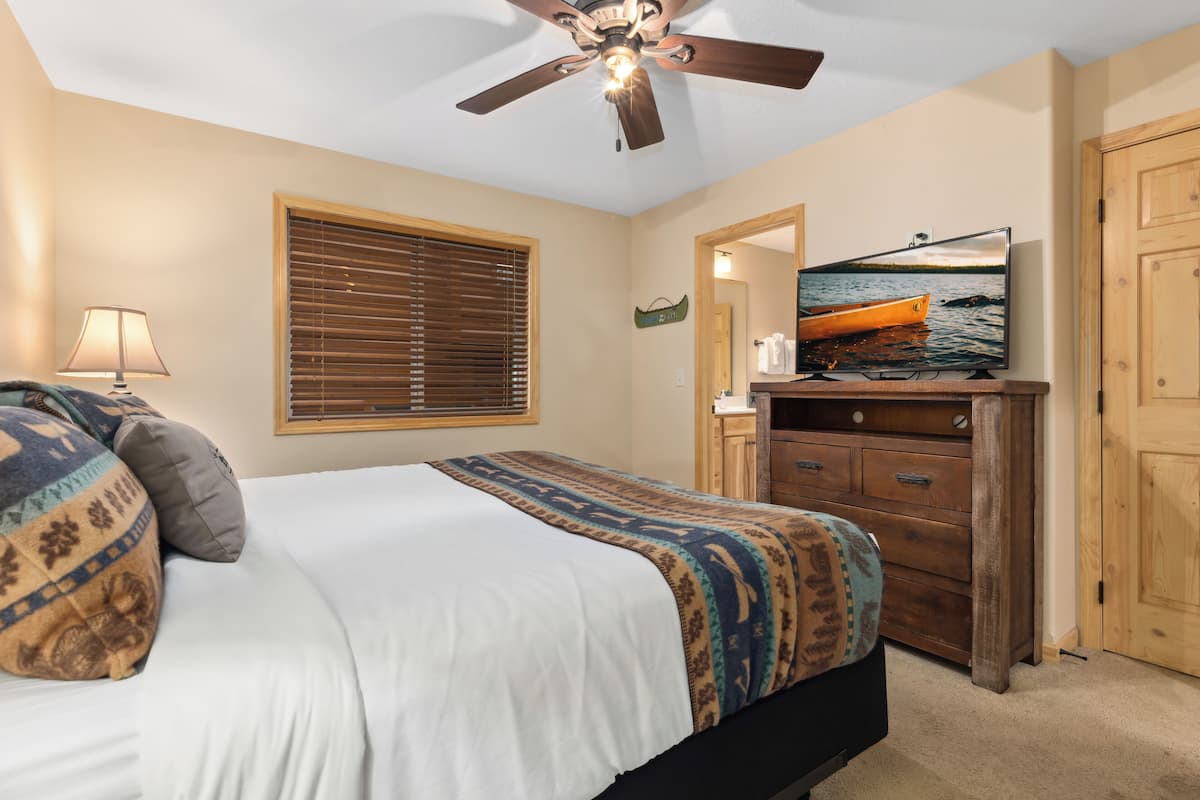 A bedroom with a bed, a ceiling fan, a television, a dresser, and a window with blinds.