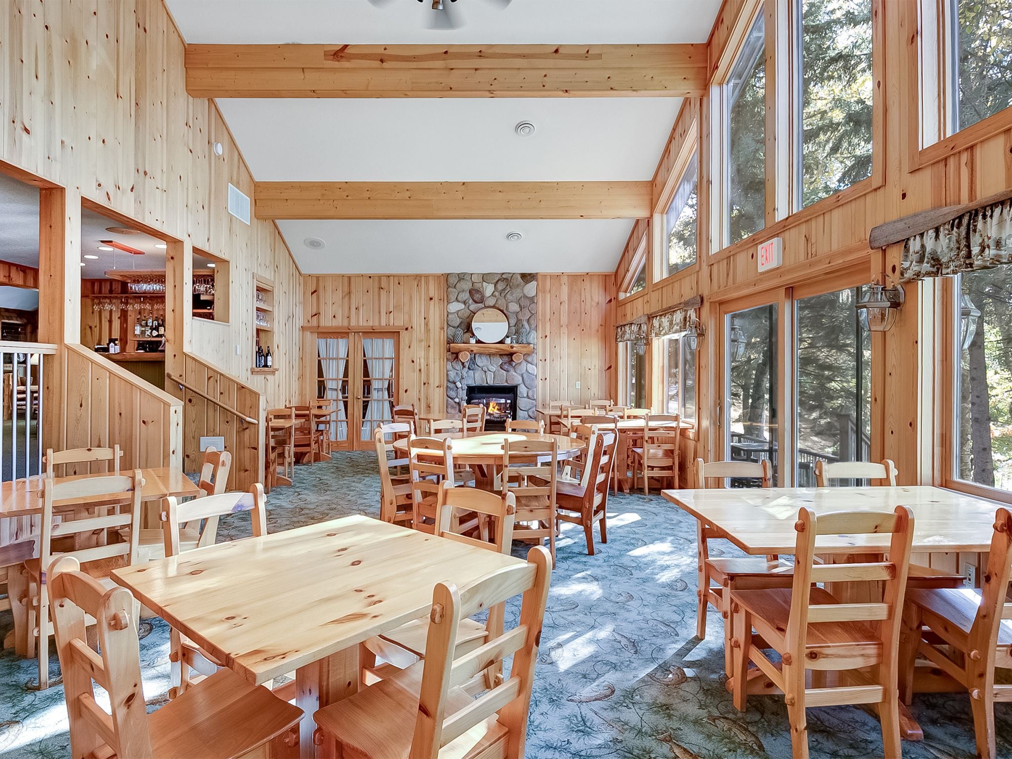 A wooden interior with multiple dining tables and chairs, a stone fireplace, and large windows.