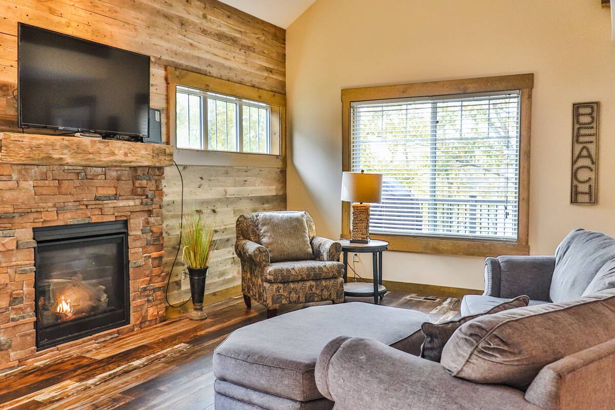 A living room with a stone fireplace, a flat-screen TV mounted above it, a comfortable armchair, a sofa, and a large window with blinds.