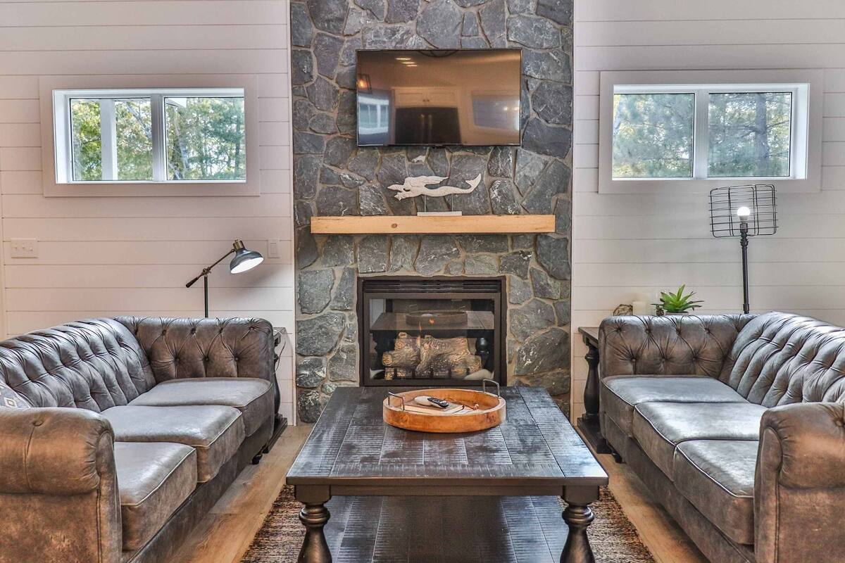 A living room with two leather sofas, a stone fireplace, a flat-screen TV, and a large window.
