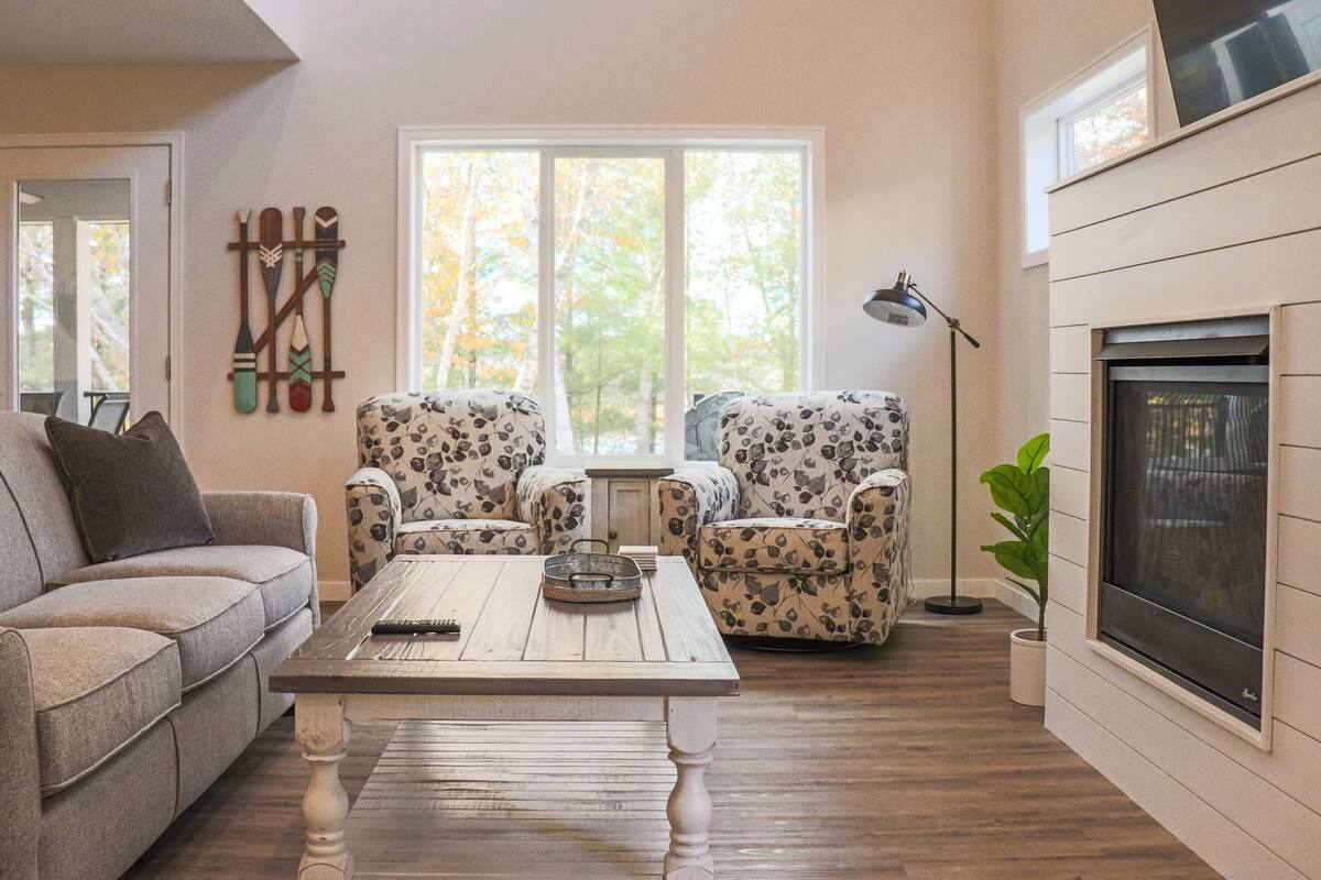 A living room with a sofa, two patterned armchairs, a coffee table, a fireplace, and a window with a view of trees.