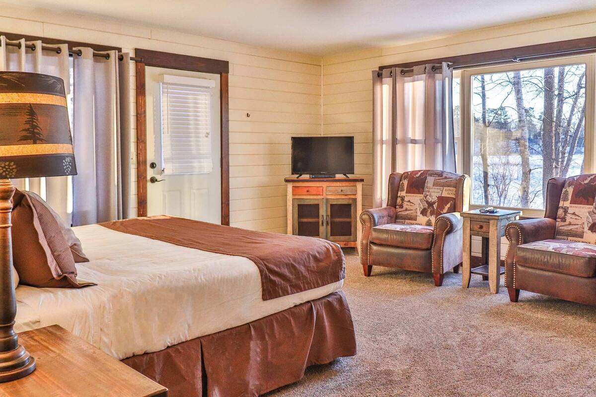 A hotel room with a bed, two armchairs, a small table, a television on a cabinet, and a window with a view of trees.
