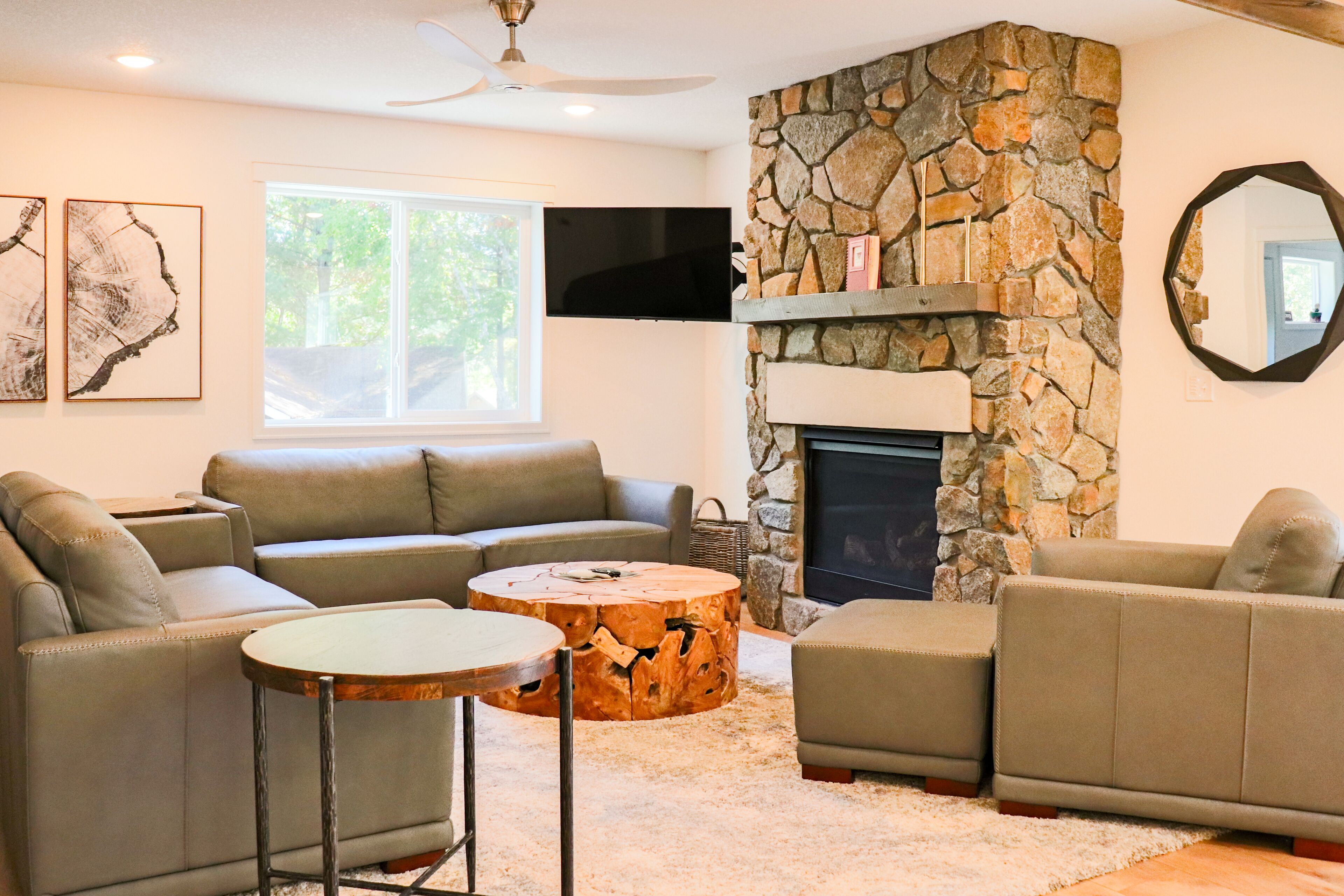 A living room with a stone fireplace, a flat-screen TV mounted above it, and a large window providing natural light.