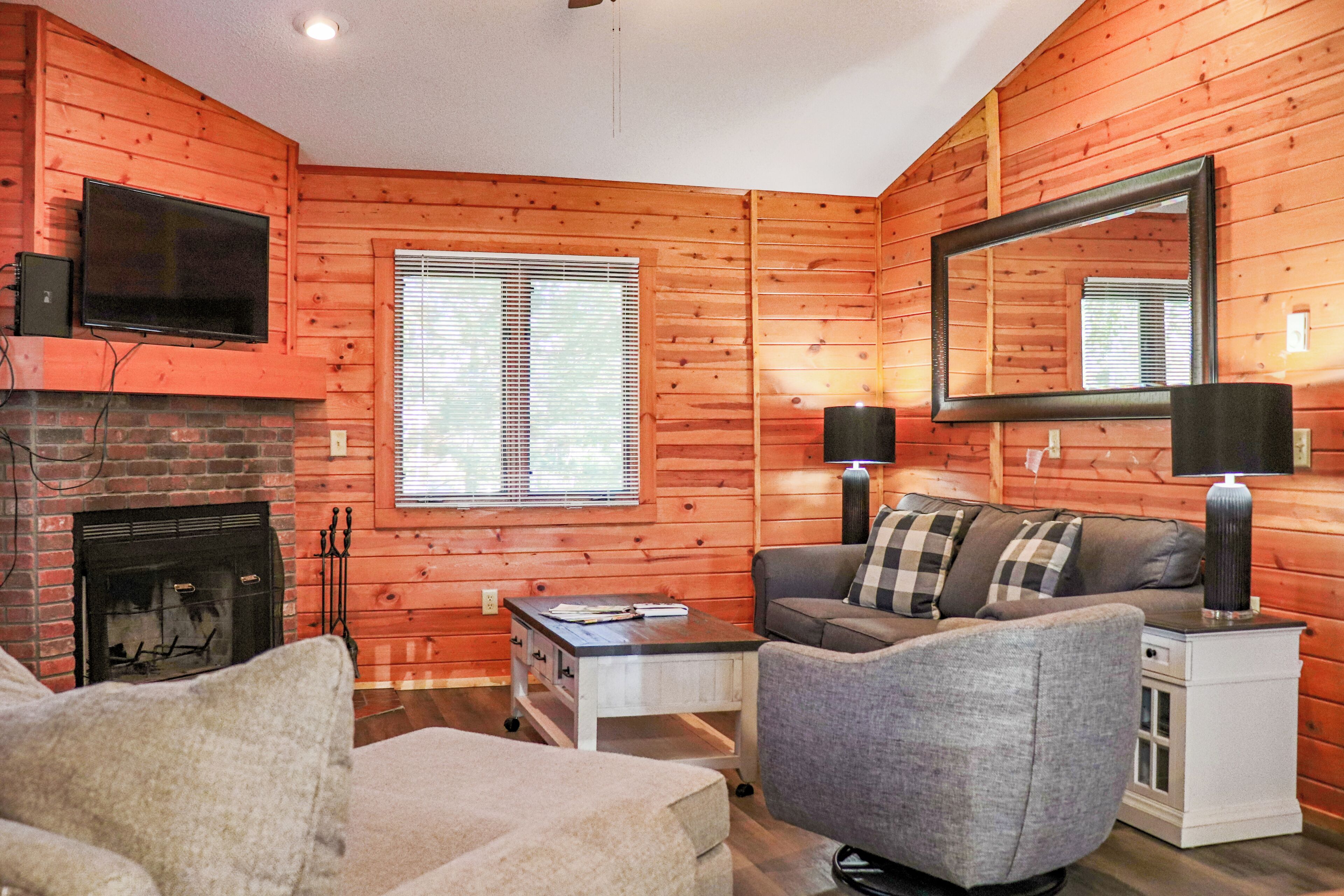 A cozy log cabin room with a fireplace, a television mounted above it, a sofa with plaid cushions, and a side table with a lamp.