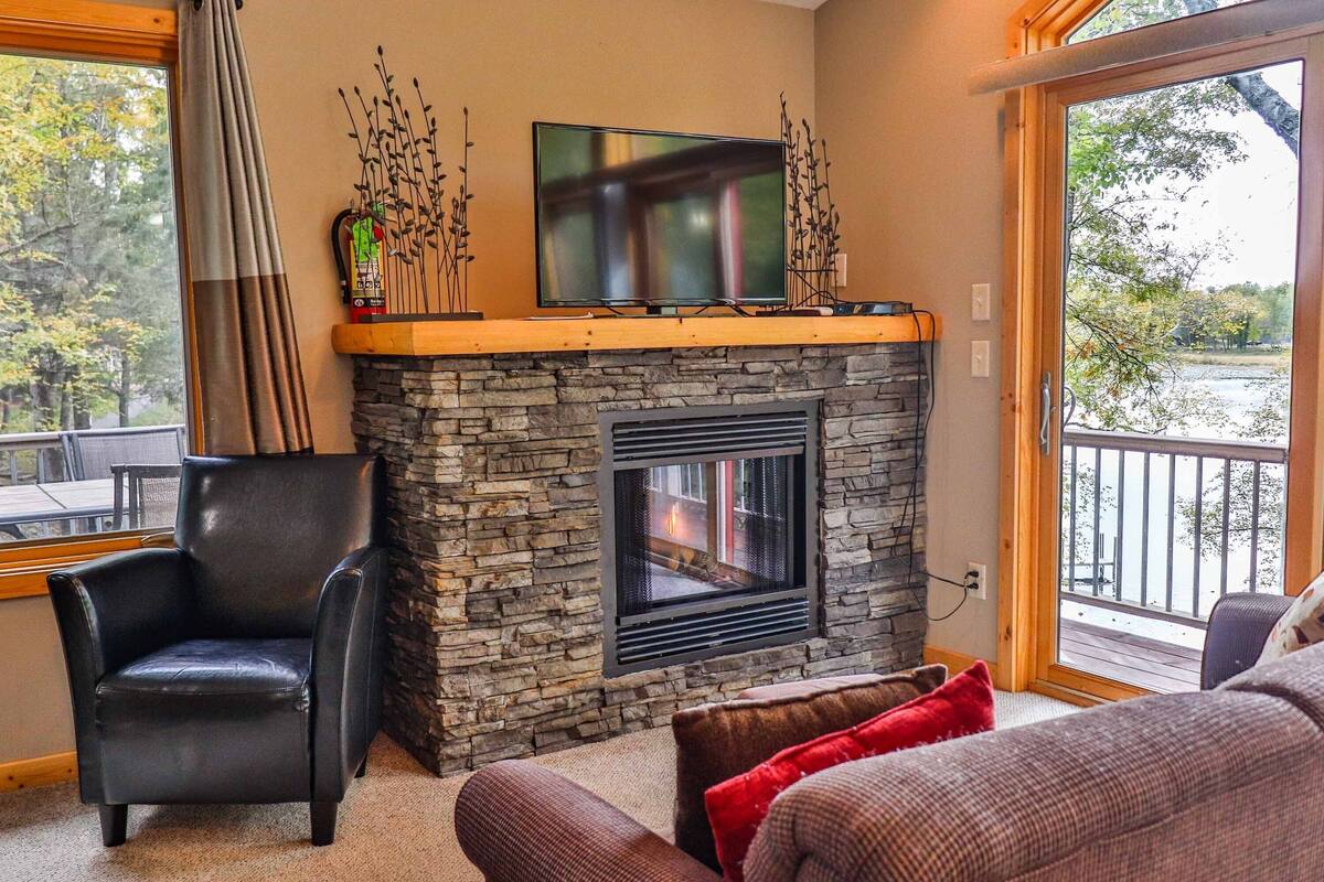 A cozy living room with a stone fireplace, a black leather chair, and a flat-screen TV on a wooden mantel.