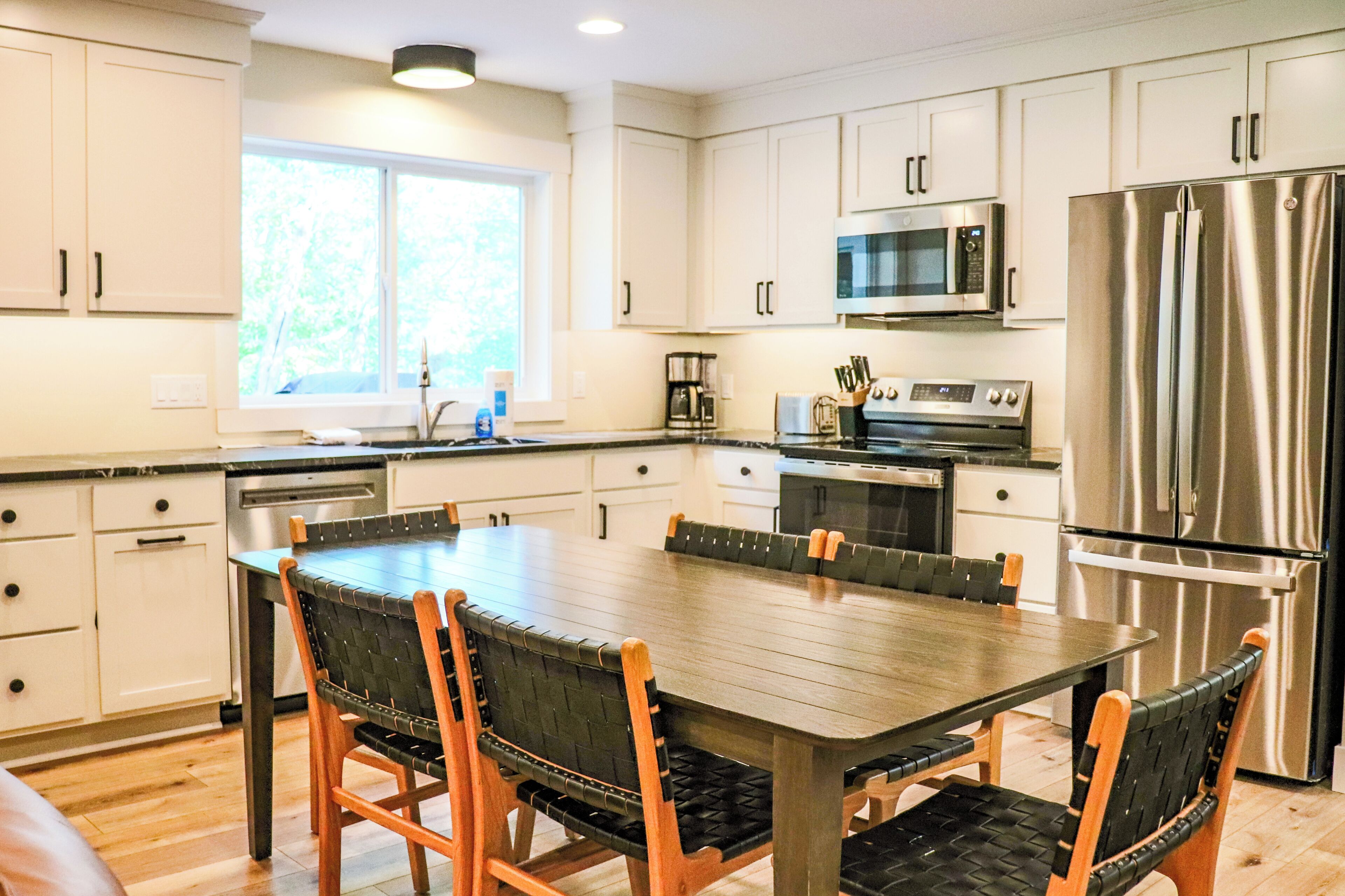 Una cocina moderna con armarios blancos, electrodomésticos de acero inoxidable, una mesa de comedor con sillas y un ventanal que proporciona luz natural.