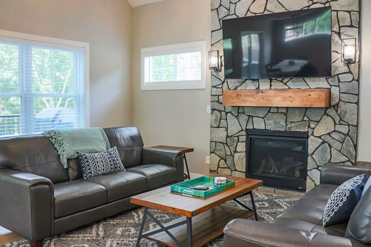 A living room with a stone fireplace, a leather sofa, a coffee table, and a flat-screen TV mounted on the wall.