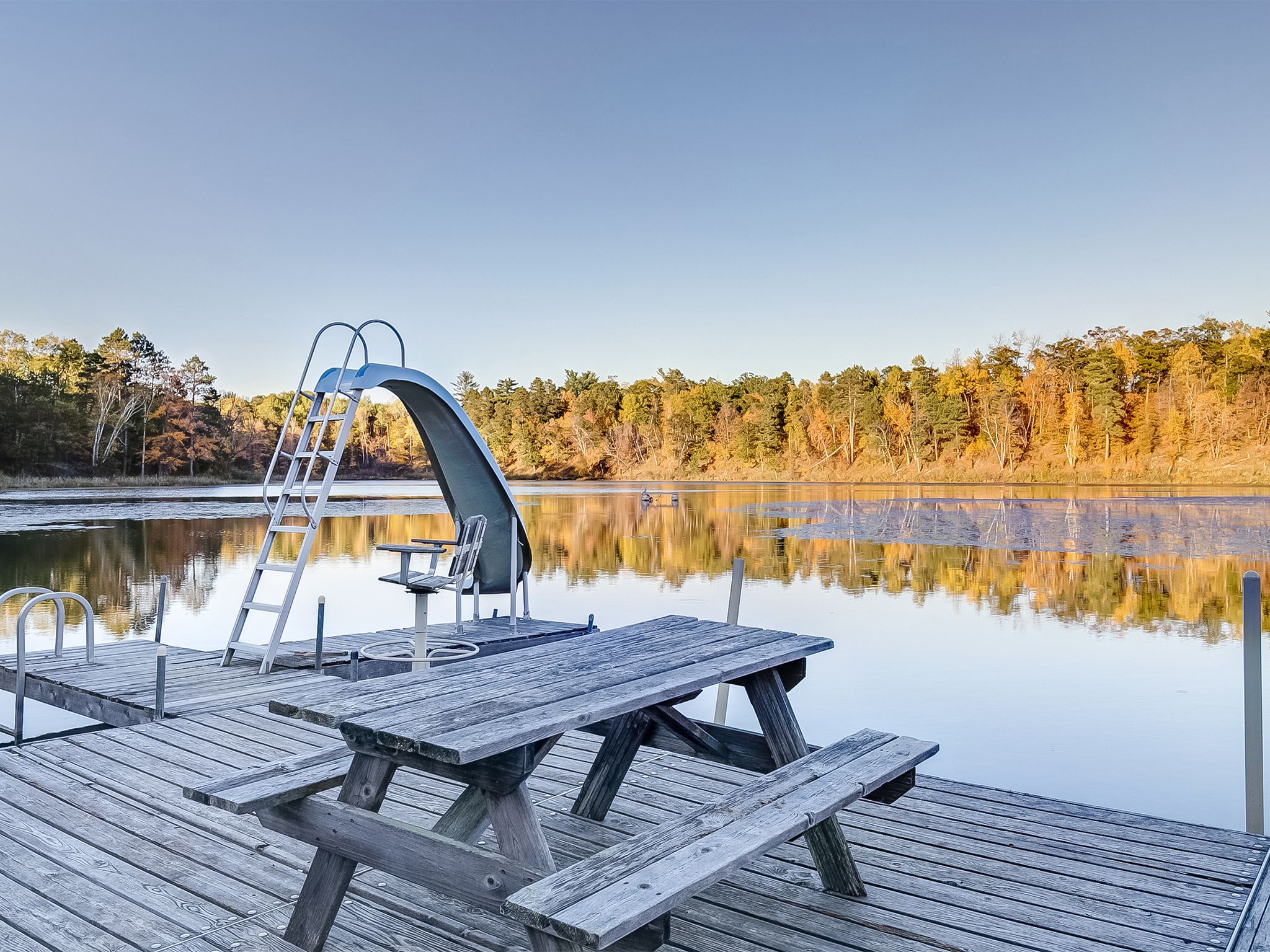A wooden dock with picnic tables and a ladder, overlooking a calm lake with trees reflecting in the water.