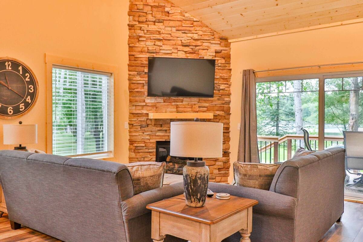 A living room with a stone fireplace, a flat-screen TV, and a view of trees through large windows.