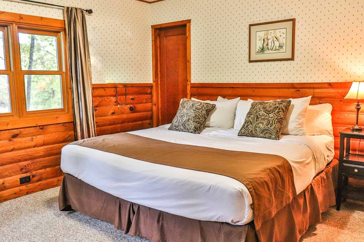 A cozy log cabin-style bedroom with a large bed, wooden walls, a window with curtains, and a framed picture on the wall.