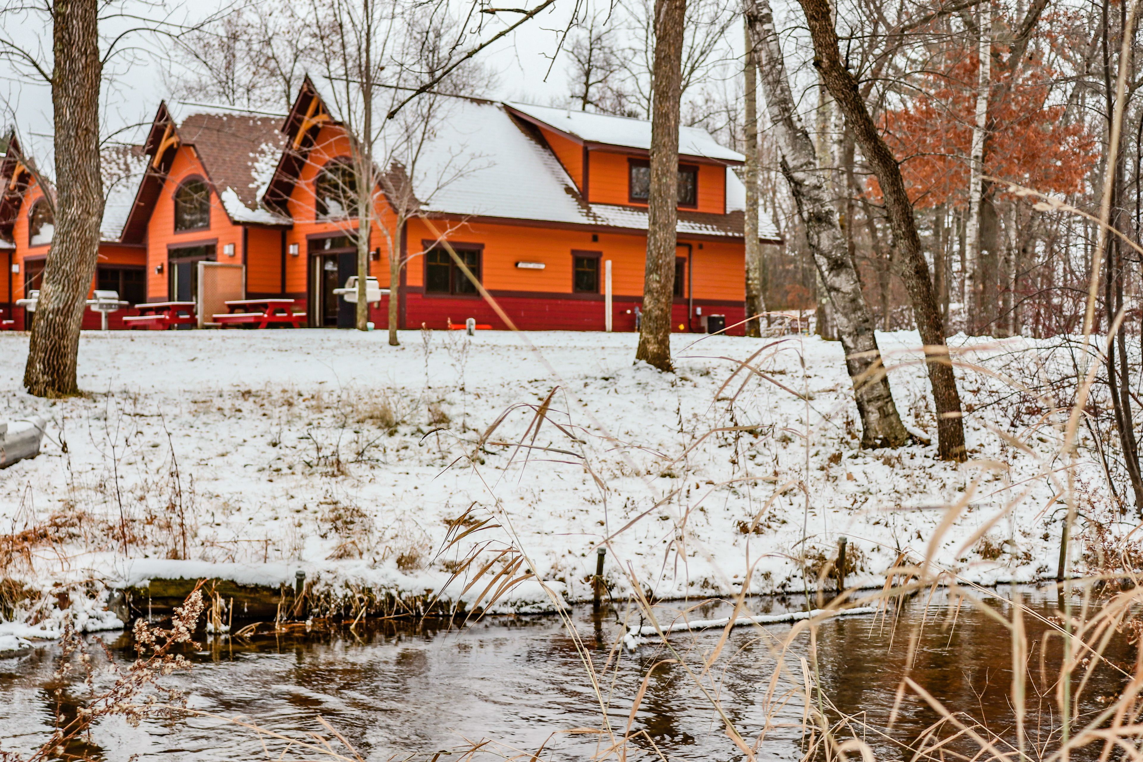 Una cabaña con fachada roja y naranja, un tejado cubierto de nieve y un pequeño arroyo en primer plano.
