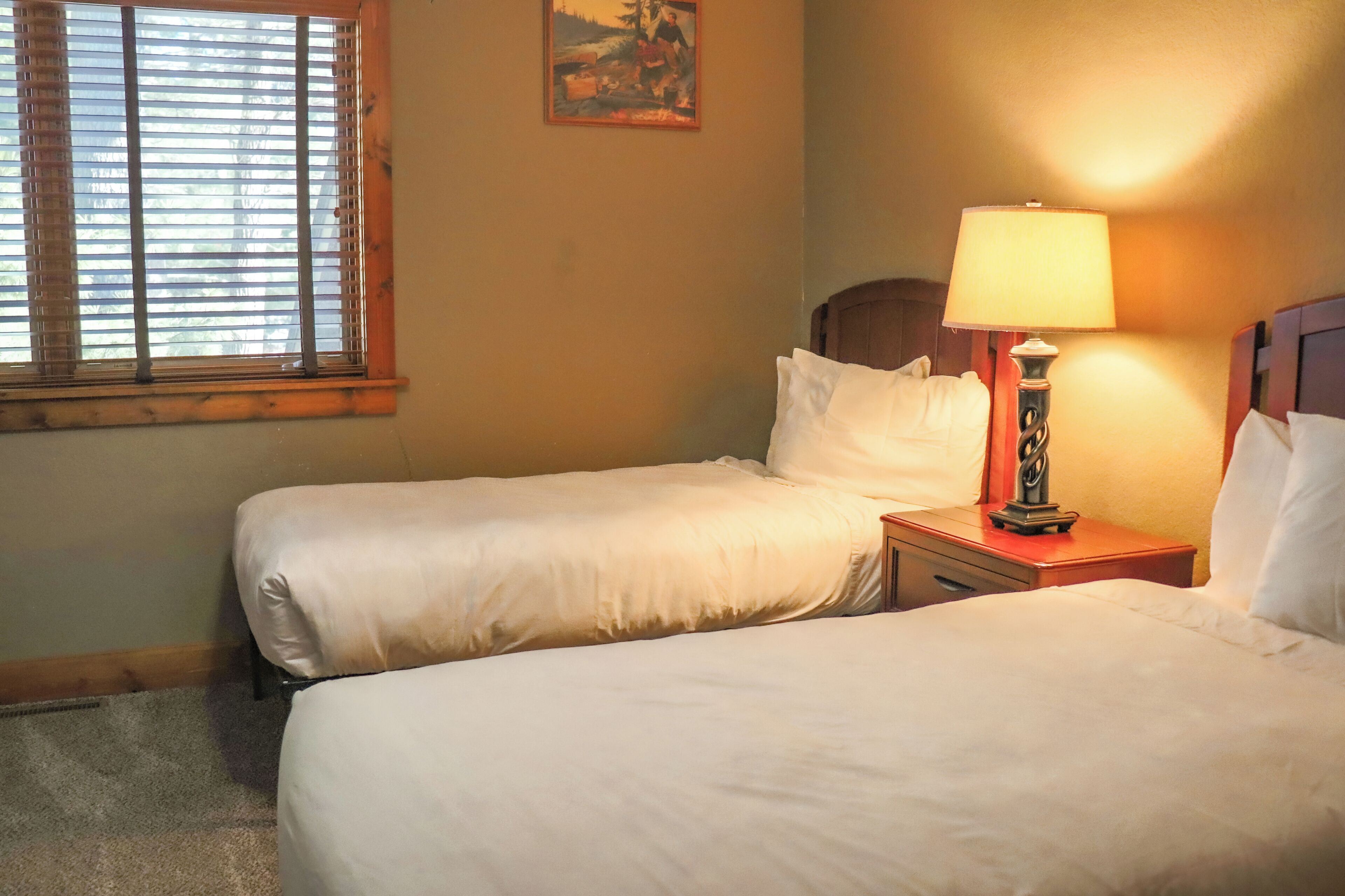 A hotel room with two beds, a nightstand with a lamp, a painting on the wall, and a window with blinds.