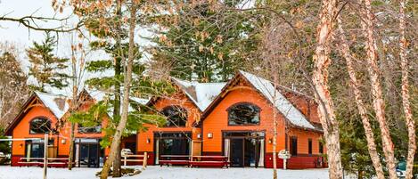 A row of wooden cabins with red roofs and large windows, surrounded by snow and trees.