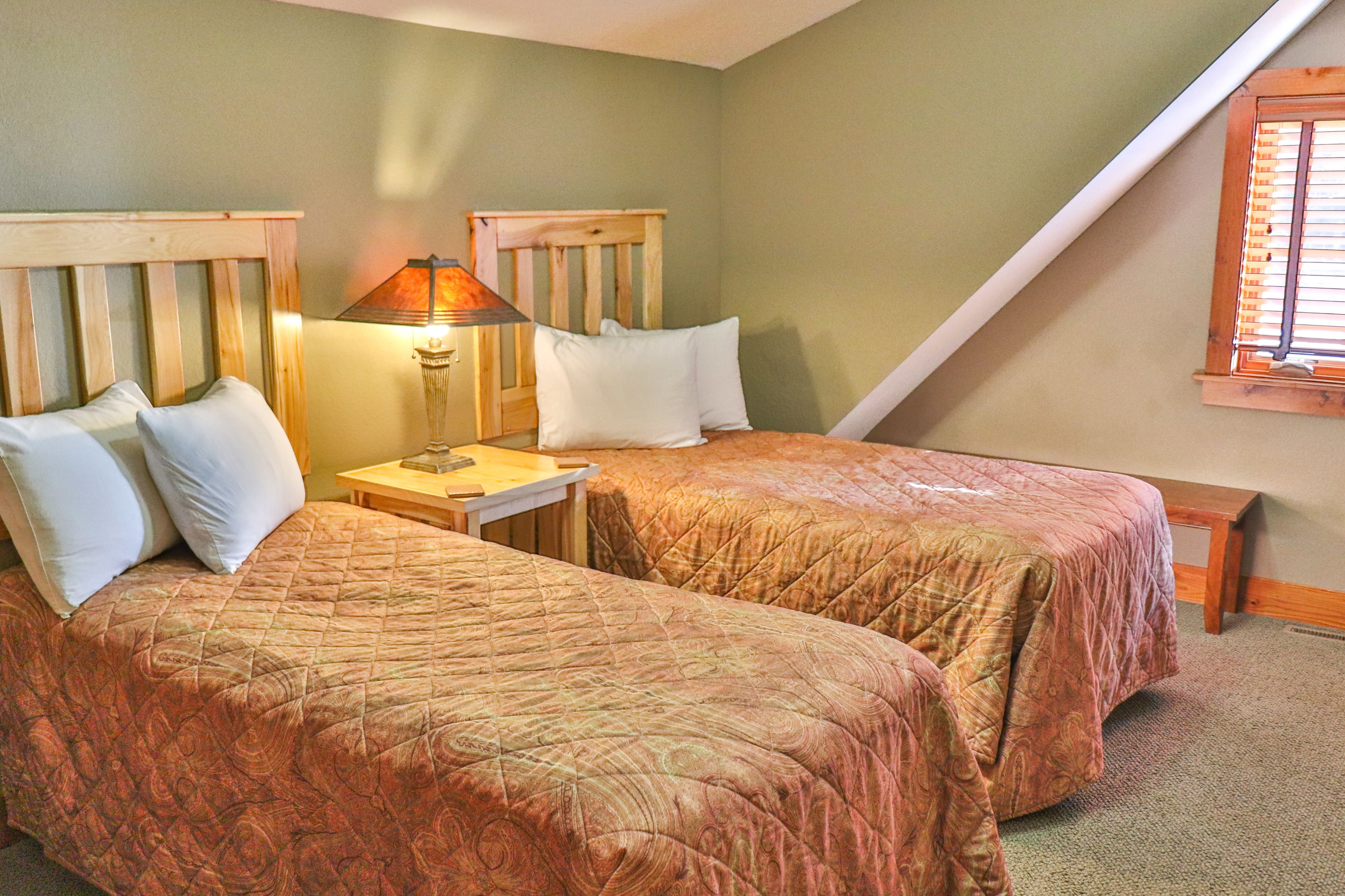 A room with two beds, each with wooden headboards and white pillows. There is a nightstand with a lamp between the beds.